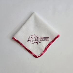 May include: White handkerchief with a burgundy crocheted edge. The word "Catherine" is embroidered in burgundy script. The handkerchief is folded in a diamond shape.