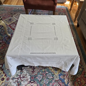 May include: A white rectangular tablecloth with embroidered geometric patterns covers a coffee table. The tablecloth has a square design in the center and decorative edges. The table is set on a patterned rug.