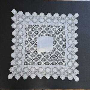 May include: A white lace doily with a square center, featuring a delicate floral pattern and a scalloped edge.