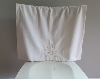 Puede incluir: Mantel de mesa de algodón blanco con un delicado diseño de encaje de ganchillo en el centro. El mantel está drapeado sobre una silla blanca.