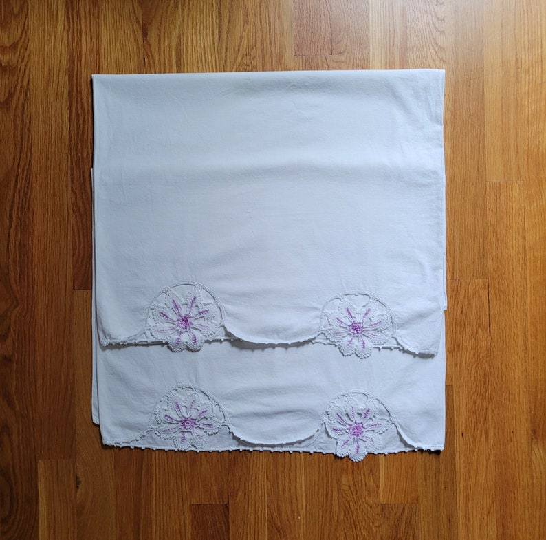 Puede incluir: Dos fundas de almohada blancas con delicado encaje y bordado floral morado. Las fundas de almohada est&aacute;n dobladas y apiladas sobre una superficie de madera.