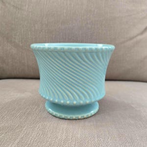 May include: A light blue ceramic planter with a flared rim and a pedestal base. The exterior features a diagonal ribbed texture across the body, accented by a delicate beaded pattern along the top rim and the base edge of the vessel.