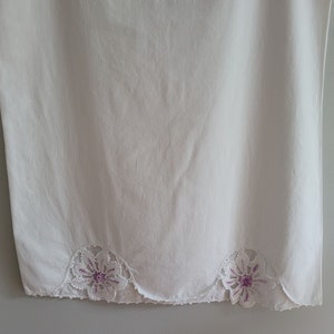 Puede incluir: Tela de algod&oacute;n blanca con un borde festoneado y dos flores bordadas de color morado.