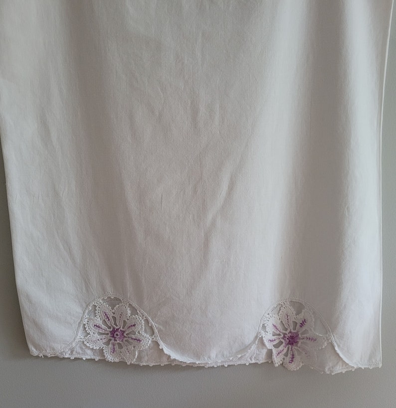 Puede incluir: Tela de algod&oacute;n blanca con un borde festoneado y dos flores bordadas de color morado.
