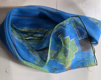 May include: A lightweight blue scarf featuring a semi-sheer fabric with a repeating geometric pattern in shades of lime green, white, and brown. The scarf has a dark green rolled hem edge and is draped in a loose, folded circular shape.