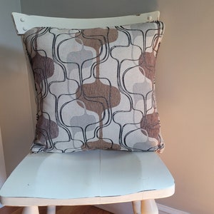 May include: A white chair with a brown and beige patterned throw pillow. The pillow has a geometric design with curved lines.