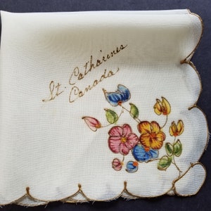 May include: White linen handkerchief with gold embroidered scalloped edges and a floral design in shades of pink, blue, yellow, and green. The handkerchief has the text "St. Catharines Canada" written in gold cursive.