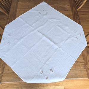 May include: White, diamond-shaped tablecloth with embroidered floral details at each point. The cloth is laid on a wooden table, showcasing its clean lines and delicate design. A classic piece for dining.