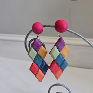 May include: A pair of colorful geometric earrings. Each earring features a pink wooden ball stud and a diamond-shaped pendant composed of various colored wooden squares, including purple, orange, green, and blue. The earrings are suspended from a metal display.