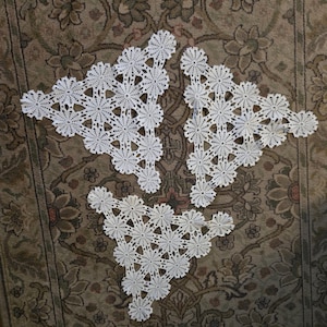 May include: Four white, handmade, crocheted doilies with a floral snowflake design are arranged on a patterned rug. The doilies are made of a delicate, open-weave material, and each has a unique shape.