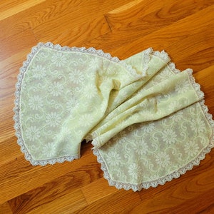 May include: A pale yellow lace tablecloth with a floral pattern. The tablecloth is folded and draped on a wooden surface.