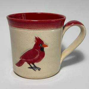 May include: A ceramic mug with a speckled off-white body and a vibrant red rim. The exterior features a raised, hand-painted red cardinal bird with a bright orange beak and black facial markings. The mug includes a sturdy, matching off-white handle.