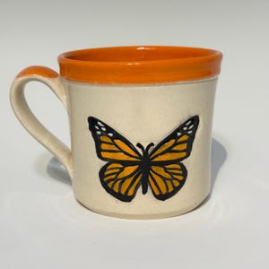 May include: A ceramic mug with a cream-colored body and an orange rim and handle. A large monarch butterfly design in orange and black is centered on the mug's front. The mug is designed for hot beverages.