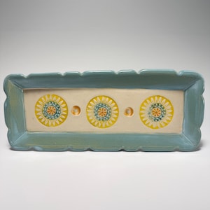 May include: A rectangular ceramic tray with a scalloped edge in a muted blue glaze. The center features a cream-colored base decorated with three yellow and orange sunflower-like floral motifs, separated by two small circular indentations.