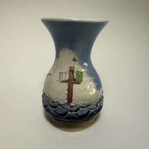 May include: A ceramic vase with a flared rim, featuring a hand-painted scene of a brown navigational marker in a blue sea under a light blue sky. The marker displays a green square sign with the number 55 and a small yellow circle icon.