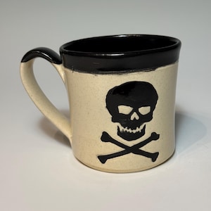 May include: A ceramic mug with a speckled off-white body and a solid black rim and handle. The front of the mug features a hand-painted black skull and crossbones design centered on the exterior surface, providing a high-contrast graphic element.