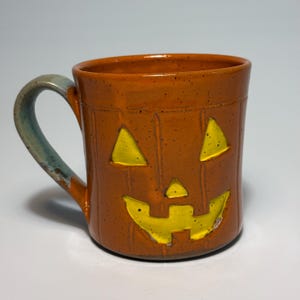May include: A ceramic mug with a speckled orange glaze and a carved jack-o'-lantern face featuring bright yellow recessed eyes, a nose, and a wide mouth. The mug has a curved handle with a blue-grey tint and vertical ridges along the exterior.