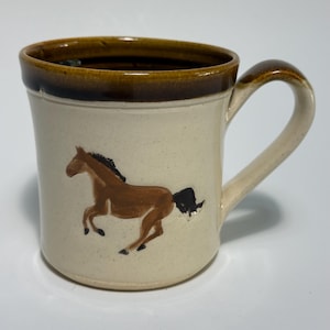 May include: A ceramic mug with a cream-colored body and a dark brown rim. The mug features a hand-painted brown horse with a black mane and tail on the side. It has a curved handle on the right side and a smooth, speckled finish throughout.