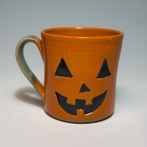May include: A ceramic mug with a glossy orange glaze and a jack-o'-lantern face featuring two triangular eyes, a small triangular nose, and a wide, jagged grin in black. The mug has a curved handle with a muted green and brown gradient finish.