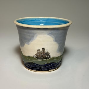 May include: Ceramic tumbler with a nautical theme. The cup features a hand-painted scene of a tall ship sailing on a wavy sea, under a cloudy sky. The rim is light blue, and the interior is a lighter shade of blue.