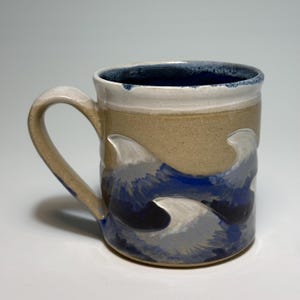 May include: A ceramic mug with a tan exterior and a deep blue interior. The lower portion of the mug features a textured, raised wave pattern in shades of blue, white, and gray, while the upper rim is finished in a smooth, off-white glaze.