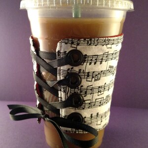 May include: A brown iced coffee drink in a clear plastic cup with a black and white fabric cup cozy with musical notes and black ribbon ties. The cozy has black buttons and is laced up the side.