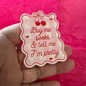May include: A pink and white gingham-patterned sign with the text "Buy me books & tell me I'm pretty." The sign has a scalloped edge, red cherries, and small hearts. The background is a bright pink.