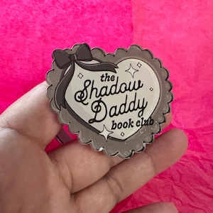 May include: A heart-shaped pin with the text "the Shadow Daddy book club" in black script. The pin has a gray scalloped border, a gray bow, and two small star accents. The background is a bright pink.
