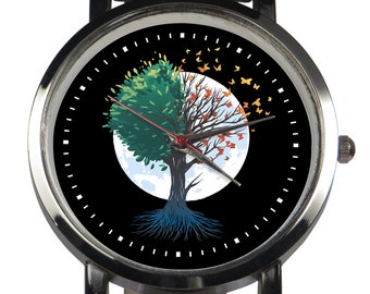 Puede incluir: Un reloj de pulsera de cuero negro con una esfera plateada. La esfera presenta un fondo negro con una luna blanca y un árbol con hojas verdes, naranjas y amarillas. El árbol tiene raíces azules y mariposas vuelan a su alrededor.