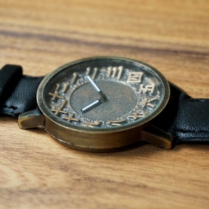 May include: A bronze-colored wristwatch with a black leather band. The watch face has Roman numerals and a textured, dark gray background.