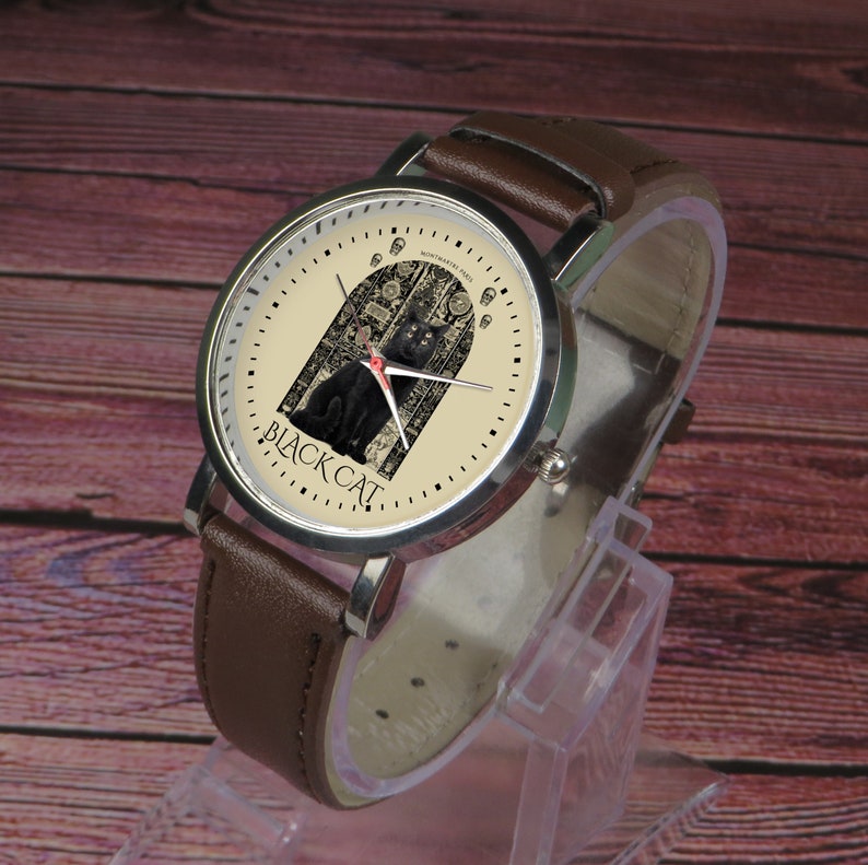May include: A brown leather wristwatch with a silver case and a cream-colored face. The face features a black cat in front of a closed door with the text "BLACK CAT" printed below the cat.