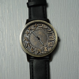 May include: A black leather wristwatch with a gold-toned face. The face has Roman numeral markings and a black hour and minute hand.
