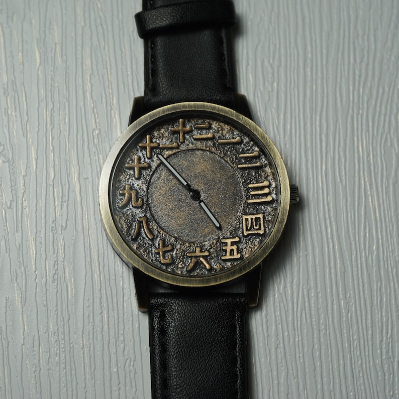 May include: A black leather wristwatch with a gold-toned face. The face has Roman numeral markings and a black hour and minute hand.