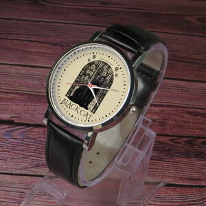 May include: A black leather wristwatch with a silver case. The watch face is cream colored with a black cat illustration and the text "BLACK CAT".