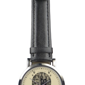 May include: A silver wristwatch with a black leather band. The watch face has a cream background with a black cat sitting in front of a detailed archway. The text "BLACK CAT" is printed below the cat. The text "MONTMARTRE PARIS" is printed above the archway.