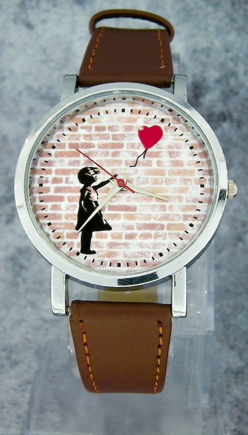 Puede incluir: Un reloj de pulsera plateado con una correa de cuero marr&oacute;n. La esfera del reloj presenta un dise&ntilde;o de pared de ladrillo con una ni&ntilde;a sosteniendo un globo rojo en forma de coraz&oacute;n.