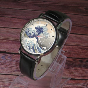 May include: A black leather wristwatch with a silver case and a white face featuring a colorful image of the Great Wave off Kanagawa by Hokusai. The watch has black hands and silver hour markers.