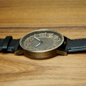 May include: A vintage-style wristwatch with a black leather band and a round, brass-colored face. The face has Roman numerals and a textured, aged look.