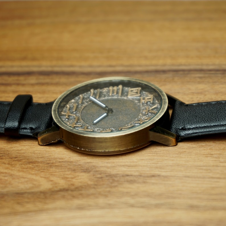 May include: A vintage-style wristwatch with a black leather band and a round, brass-colored face. The face has Roman numerals and a textured, aged look.