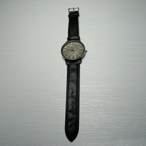 May include: A black leather wristwatch with a round, silver-toned face. The face has black Roman numeral markings and a black second hand.