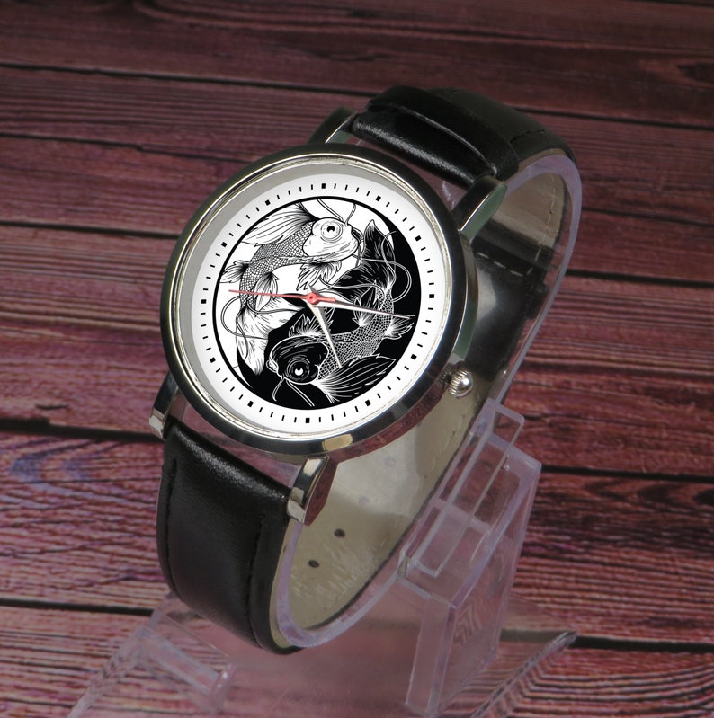 May include: A black leather wristwatch with a silver face and a yin yang design featuring two black and white fish. The watch is on a clear display stand.