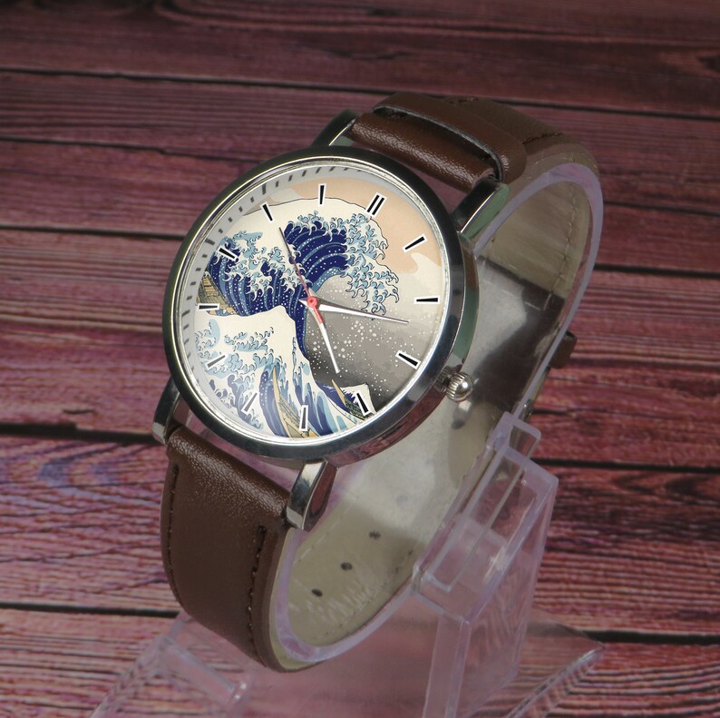 May include: A brown leather wristwatch with a silver case and a white face. The face features a colorful image of a wave, reminiscent of the Great Wave off Kanagawa by Hokusai.