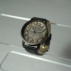 May include: A black leather wristwatch with a round, brass-colored face. The face has Japanese numerals for the hours, and the hands are white.