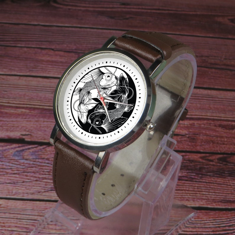 May include: A brown leather wristwatch with a silver face and black and white yin yang design featuring two fish.