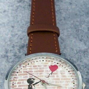Puede incluir: Un reloj de pulsera plateado con una correa de cuero marr&oacute;n. La esfera del reloj presenta una pared de ladrillo con una ni&ntilde;a sosteniendo un globo rojo en forma de coraz&oacute;n.