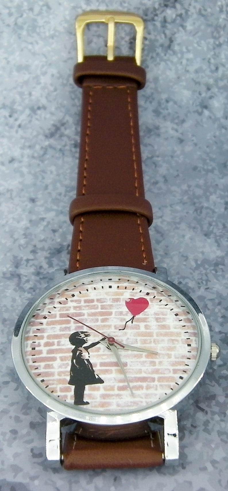 Puede incluir: Un reloj de pulsera plateado con una correa de cuero marr&oacute;n. La esfera del reloj presenta una pared de ladrillo con una ni&ntilde;a sosteniendo un globo rojo en forma de coraz&oacute;n.