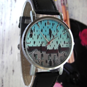 May include: A silver wristwatch with a black leather band. The watch face features a blue and white print of a painting with figures on a rooftop.