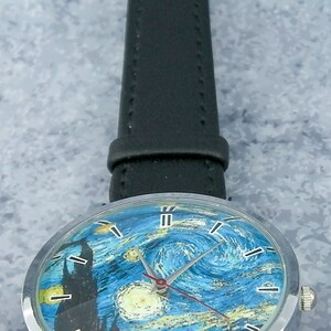 May include: A silver wristwatch with a black leather band. The watch face features a blue and yellow painting of a starry night.