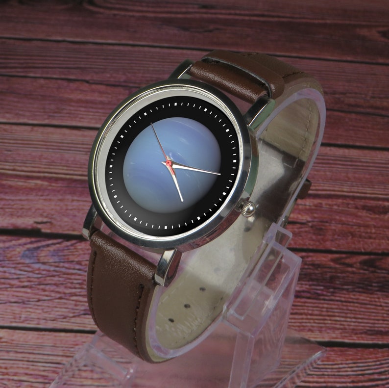 May include: A silver watch with a brown leather strap. The watch face has a black background with a white circle in the centre. The circle has a blue and white design that resembles a planet.