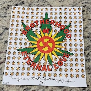 Peut inclure: Une feuille de papier avec un motif répété de petits motifs floraux orange et verts. Le centre présente un grand dessin avec les mots "BROTHERHOOD OF ETERNAL LOVE" et un symbole en forme de soleil. Signé par Robert Ackerly, Mike Hynson et Eddie Dodge.